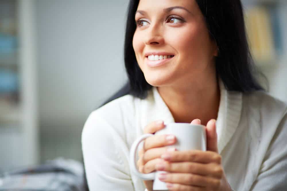 A Woman Holding A Cup Of Coffee And Smiling - Showing The Effects Of Coffee On Teeth.