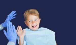 Smiling young boy in a dental bib raising his hand toward a gloved dental team member for a high five.